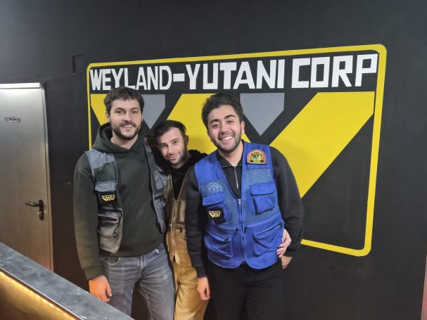 Tres personas posan frente a un fondo de la corporación Weyland-Yutani.
